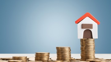 Une petite maison avec un toit rouge est perchée sur une haute pile de pièces de monnaie, symbolisant la croissance des investissements immobiliers.