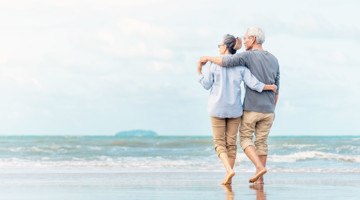 Een ouder paar wandelt met hun armen om elkaar heen langs het strand, met de zee op de achtergrond.