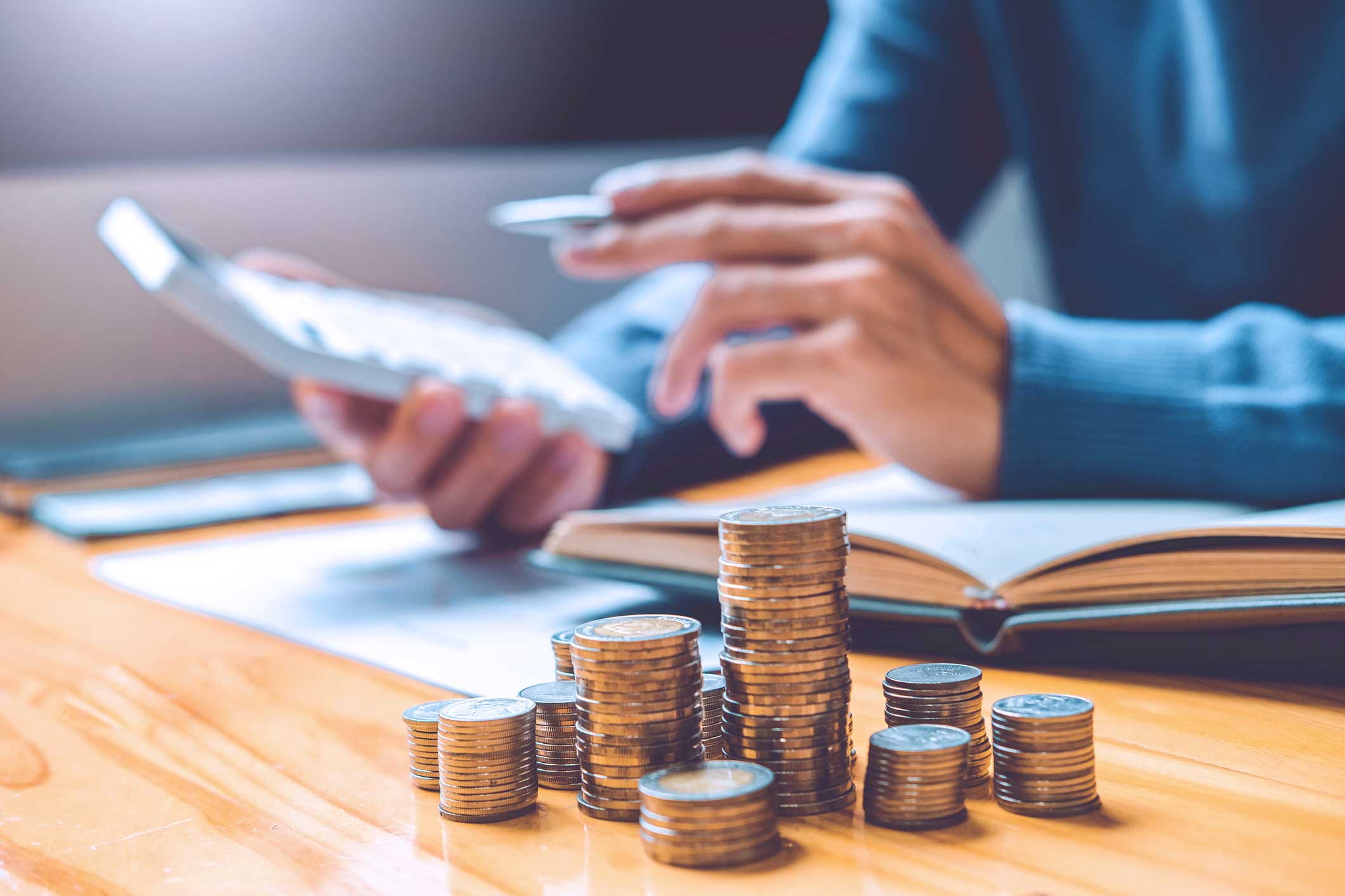 Une main avec des pièces de monnaie et une calculatrice, entourée de piles de pièces et d'un livre ouvert sur une table en bois.