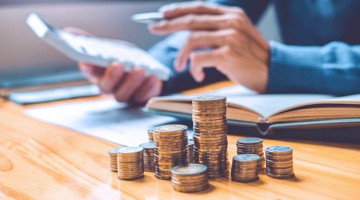 Une main avec des pièces de monnaie et une calculatrice, entourée de piles de pièces et d'un livre ouvert sur une table en bois.