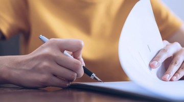 Une main tient un stylo et feuillette une pile de documents.