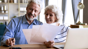 Un couple âgé consulte des documents à une table avec un ordinateur portable et des notes.
