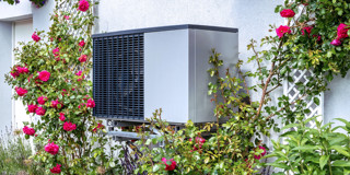 Een aan de muur geïnstalleerde luchtwarmtepomp, omringd door bloeiende rozen en groene planten.