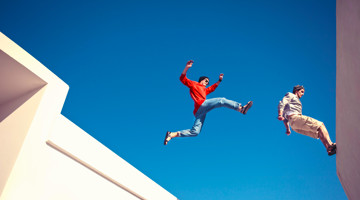 Twee personen springen tussen witte gebouwen tegen een heldere blauwe lucht; één persoon in een rode trui en jeans zweeft midden in de sprong, terwijl een andere persoon in lichte kleding net van een rand afspringt.