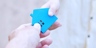 Een hand geeft een blauwe sleutelhanger in de vorm van een huisje aan een andere hand, wat woningoverdracht of nieuwe mogelijkheden symboliseert.