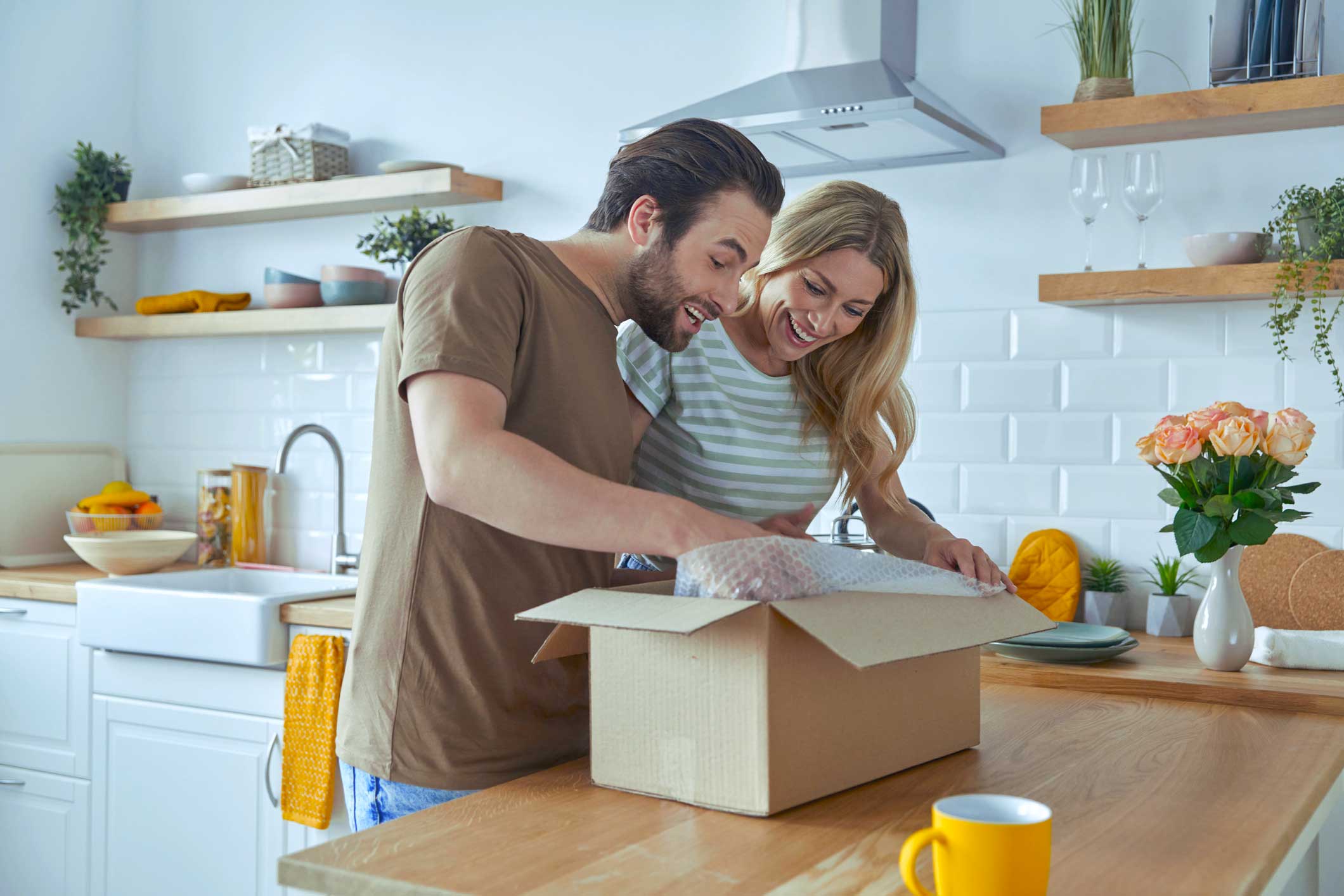 Een man en vrouw openen samen een kartonnen doos in een moderne keuken, omringd door planten en keukenaccessoires.