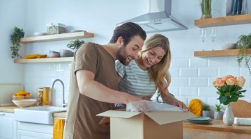 Een man en vrouw openen samen een kartonnen doos in een moderne keuken, omringd door planten en keukenaccessoires.