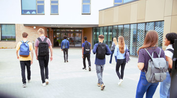 Een groep studenten met rugzakken wandelt naar de ingang van een modern schoolgebouw.