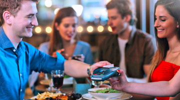 Une personne dans un restaurant paie avec un terminal de carte bancaire pendant que d'autres sont à table et utilisent leurs téléphones.