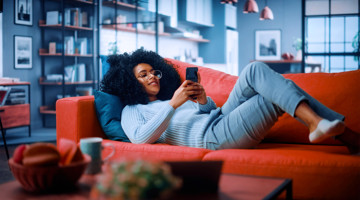 Vrouw met krullend haar in een lichte trui ligt op een rode bank en kijkt naar haar smartphone.