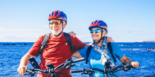 Twee fietsers met helmen staan naast hun fietsen aan de kust, met een heldere blauwe lucht en zee op de achtergrond.