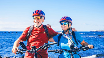 Twee fietsers met helmen staan naast hun fietsen aan de kust, met een heldere blauwe lucht en zee op de achtergrond.