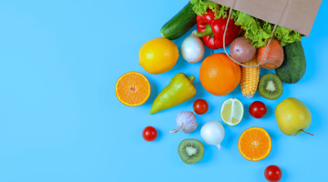 Des fruits et légumes frais, notamment des poivrons, des oranges, des concombres et des tomates, sont disposés sur un fond bleu, avec une boîte ouverte en arrière-plan.