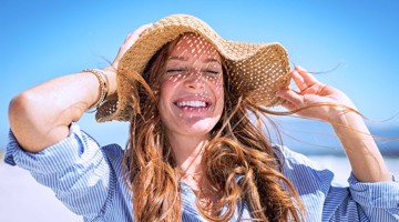 Femme avec un chapeau de paille, debout sur la plage, avec un ciel bleu clair en arrière-plan.