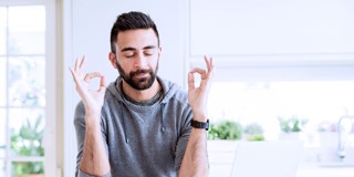 Homme en hoodie grise faisant un geste de main 'ok' avec les deux mains, assis à un bureau avec un verre d'eau.