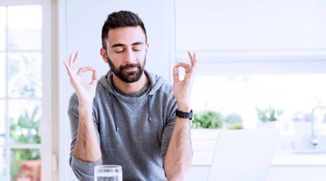 Homme en hoodie grise faisant un geste de main 'ok' avec les deux mains, assis à un bureau avec un verre d'eau.