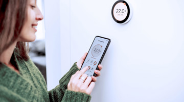 Une femme utilise son smartphone pour régler un thermostat intelligent, affichant une température de 22°C.