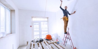 Un homme peint un mur blanc en étant sur une échelle ; des planches en bois et un casque de chantier sont posés sur le sol.