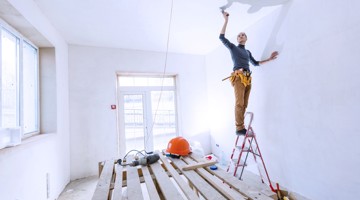 Un homme peint un mur blanc en étant sur une échelle ; des planches en bois et un casque de chantier sont posés sur le sol.