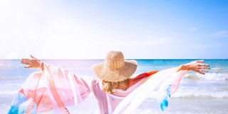 Een vrouw met een strohoed staat met open armen aan de kust, met de zee en een blauwe lucht op de achtergrond.