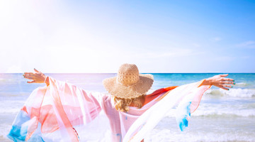 Een vrouw met een strohoed staat met open armen aan de kust, met de zee en een blauwe lucht op de achtergrond.