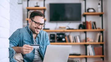 Un homme en veste en jean tient une carte de crédit et regarde un ordinateur portable dans un salon moderne, avec des étagères de livres en arrière-plan.