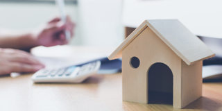 Modèle en bois d'une maison sur un bureau, avec en arrière-plan une main prenant des notes à côté d'une calculatrice.