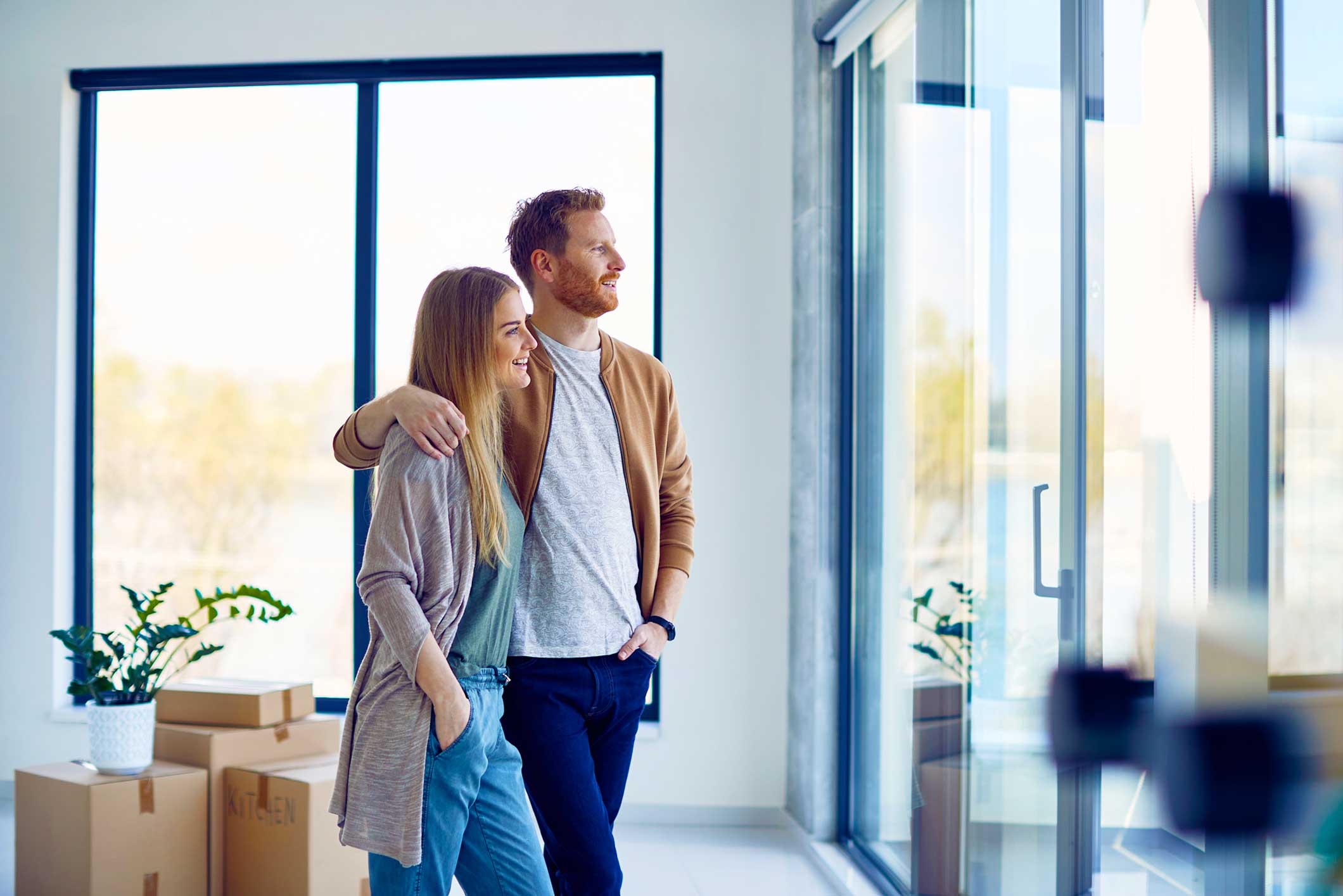 Jong stel staat naast elkaar in een moderne, lichte woonkamer met grote ramen en planten op de achtergrond.