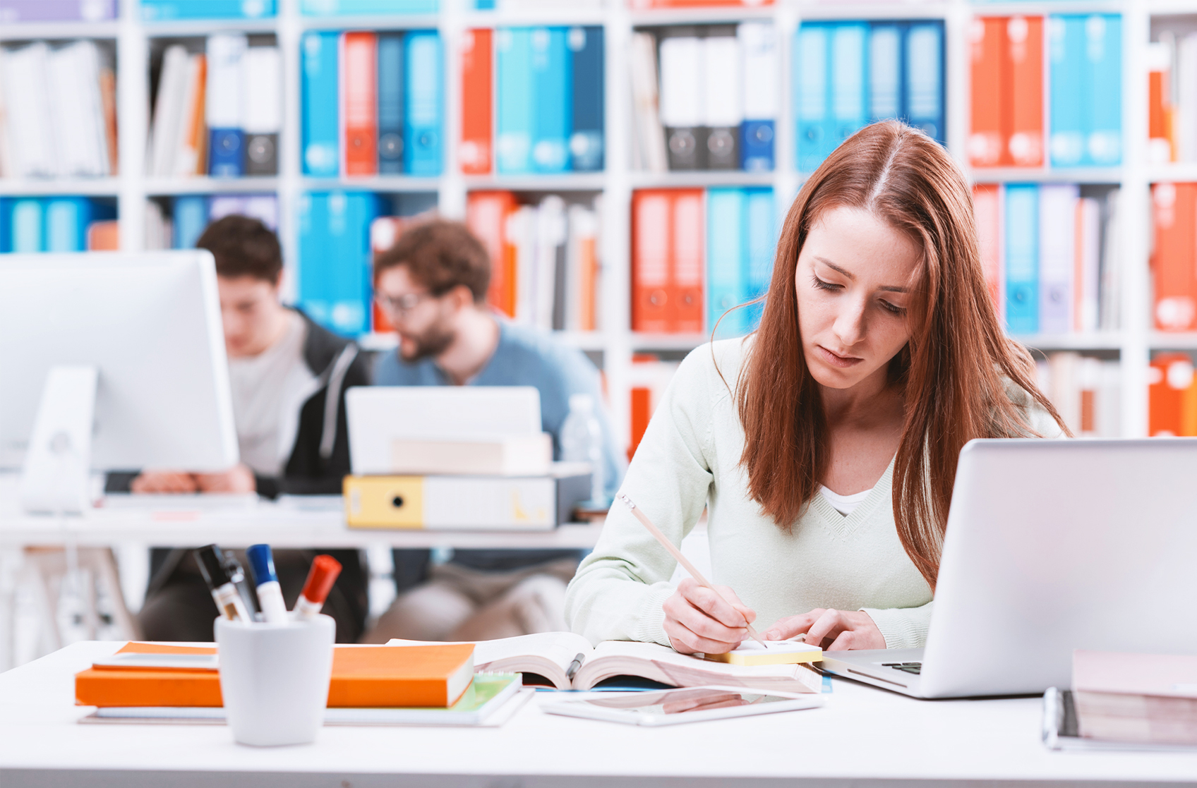 Een studente maakt aantekeningen aan haar bureau met een laptop, omringd door boeken in een bibliotheek.