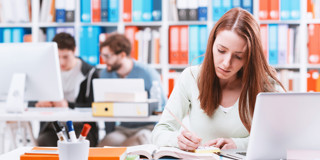 Een studente maakt aantekeningen aan haar bureau met een laptop, omringd door boeken in een bibliotheek.