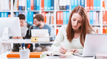 Een studente maakt aantekeningen aan haar bureau met een laptop, omringd door boeken in een bibliotheek.