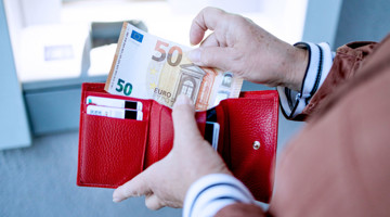 Een hand houdt een vijftig euro biljet boven een open rode portemonnee met zichtbare kaarten.