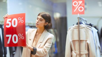 Femme présentant des réductions de 50 % et 70 % sur des panneaux rouges dans un magasin de vêtements.