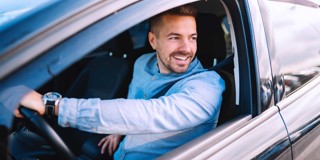 Een persoon in een auto, gekleed in een blauwe trui en een polshorloge, zit op de bestuurdersstoel met de arm op de raamlijn.