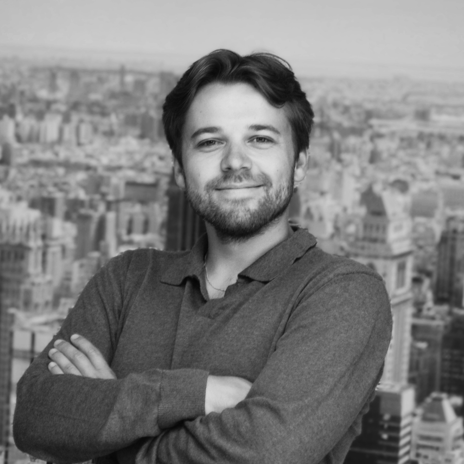 Een man in een grijs overhemd staat met zijn armen over elkaar, tegen een zwart-witte stadse achtergrond.