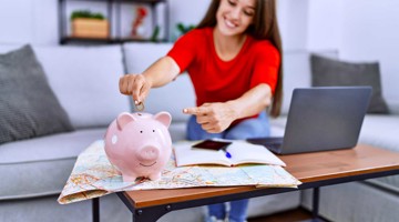 Femme en chemise rouge met une pièce dans une tirelire rose sur une table avec une carte et un ordinateur portable.