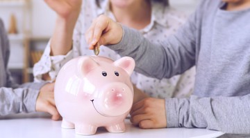Des enfants mettent des pièces de monnaie dans une tirelire rose en forme de cochon, symbolisant l'épargne et l'éducation financière.