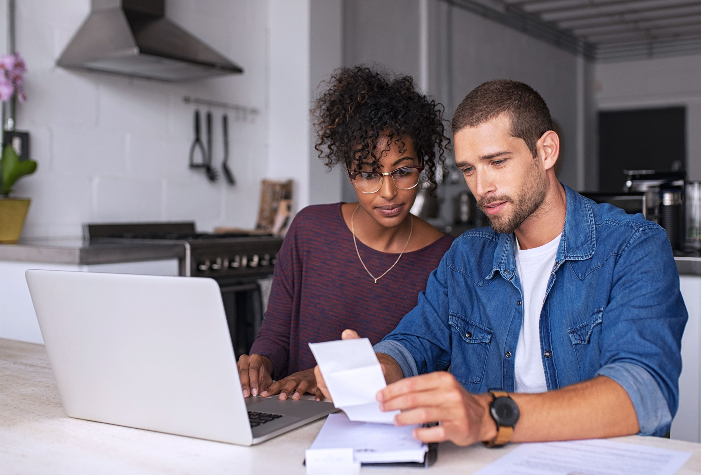 Een vrouw en een man bekijken samen een bon terwijl ze naar een laptop in een moderne keuken kijken.