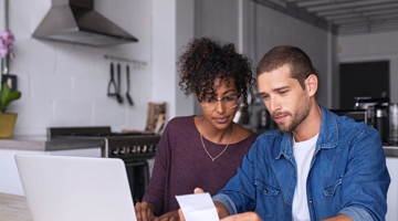 Een vrouw en een man bekijken samen een bon terwijl ze naar een laptop in een moderne keuken kijken.