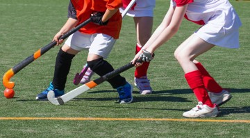 Drie meisjes in sportkleding spelen hockey op een grasveld en proberen de bal met hun hockeysticks te raken.