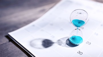 Sablier avec des grains de sable bleus sur un calendrier blanc, symbolisant la pression du temps et les délais.